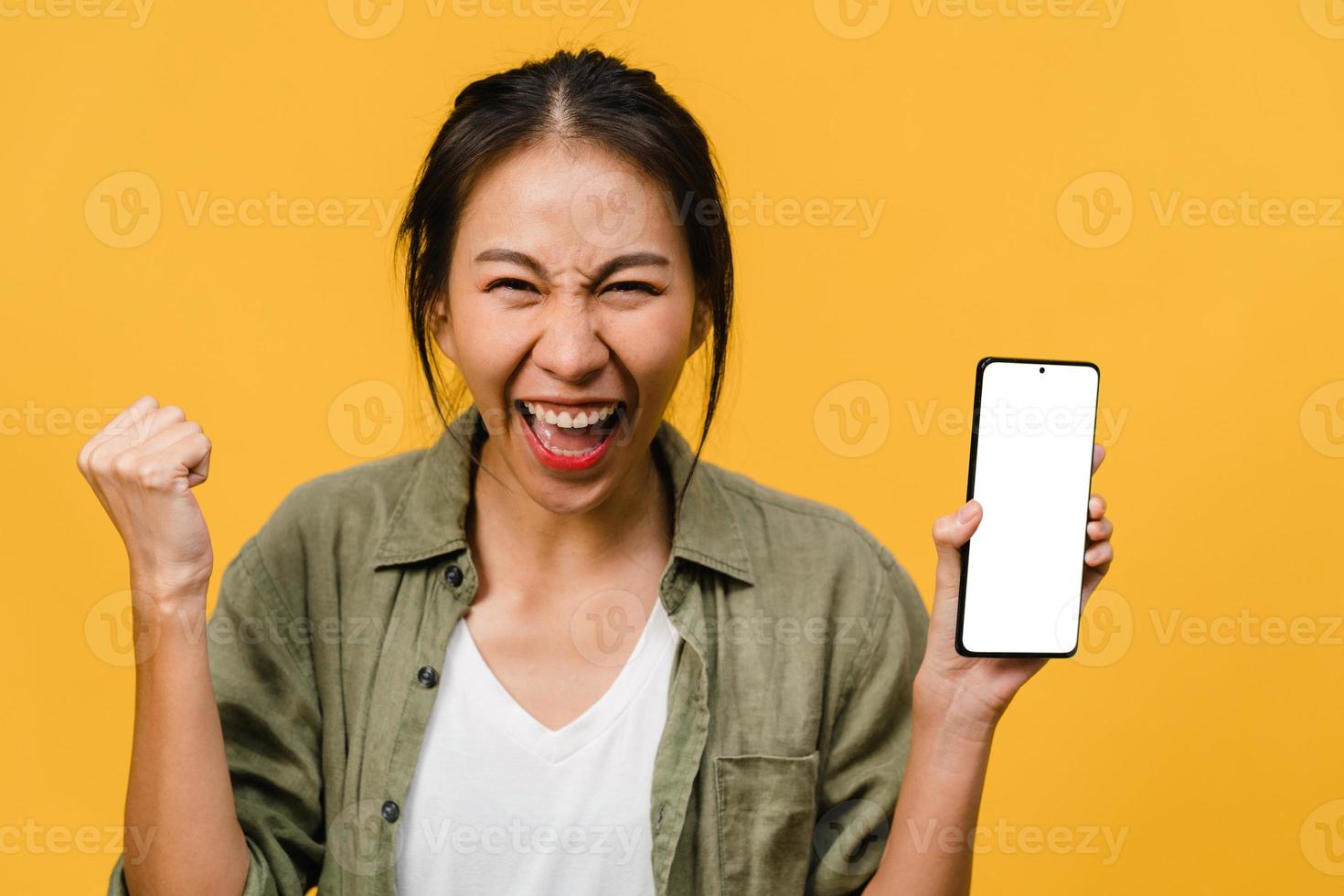 jonge aziatische dame toont een leeg smartphonescherm met positieve uitdrukking, glimlacht breed, gekleed in casual kleding en voelt zich gelukkig op gele achtergrond. mobiele telefoon met wit scherm in vrouwelijke hand. foto