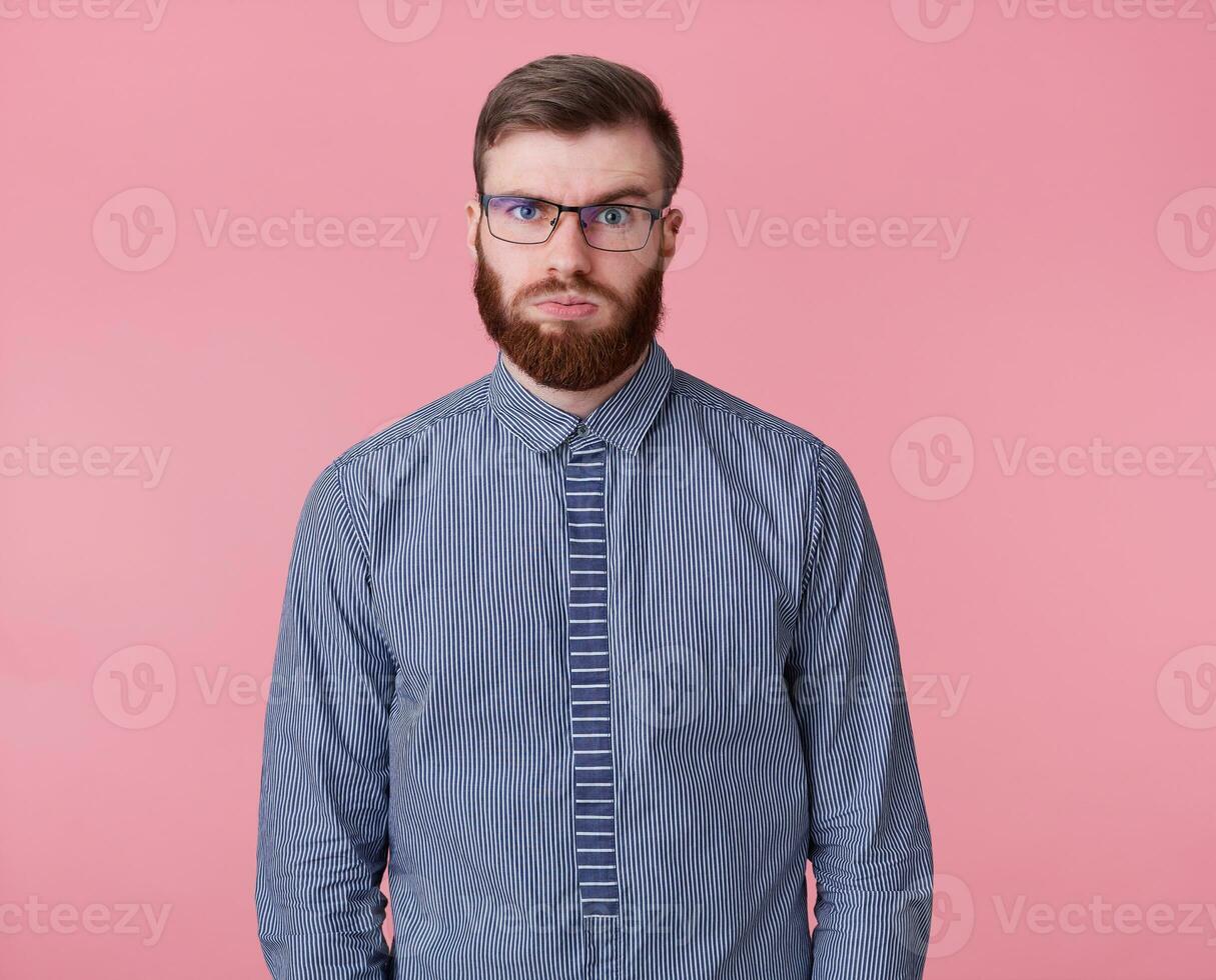 jong moe van de werk rood gebaard vent met bril en een gestreept overhemd looks in de afstand, denkt hoe veel meer projecten naar dichtbij en of hij zullen hebben tijd, geïsoleerd over- roze achtergrond. foto