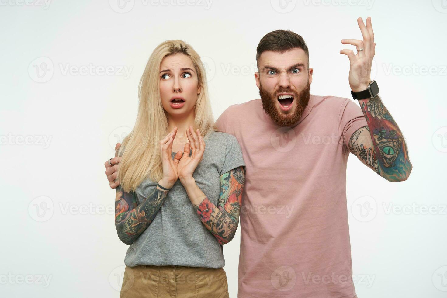 geërgerd jong gebaard bruin haar getatoeëerd vent fronsen boos zijn gezicht en verhogen emotioneel hand- terwijl knuffelen zijn verward blond vriendin, geïsoleerd over- wit achtergrond foto