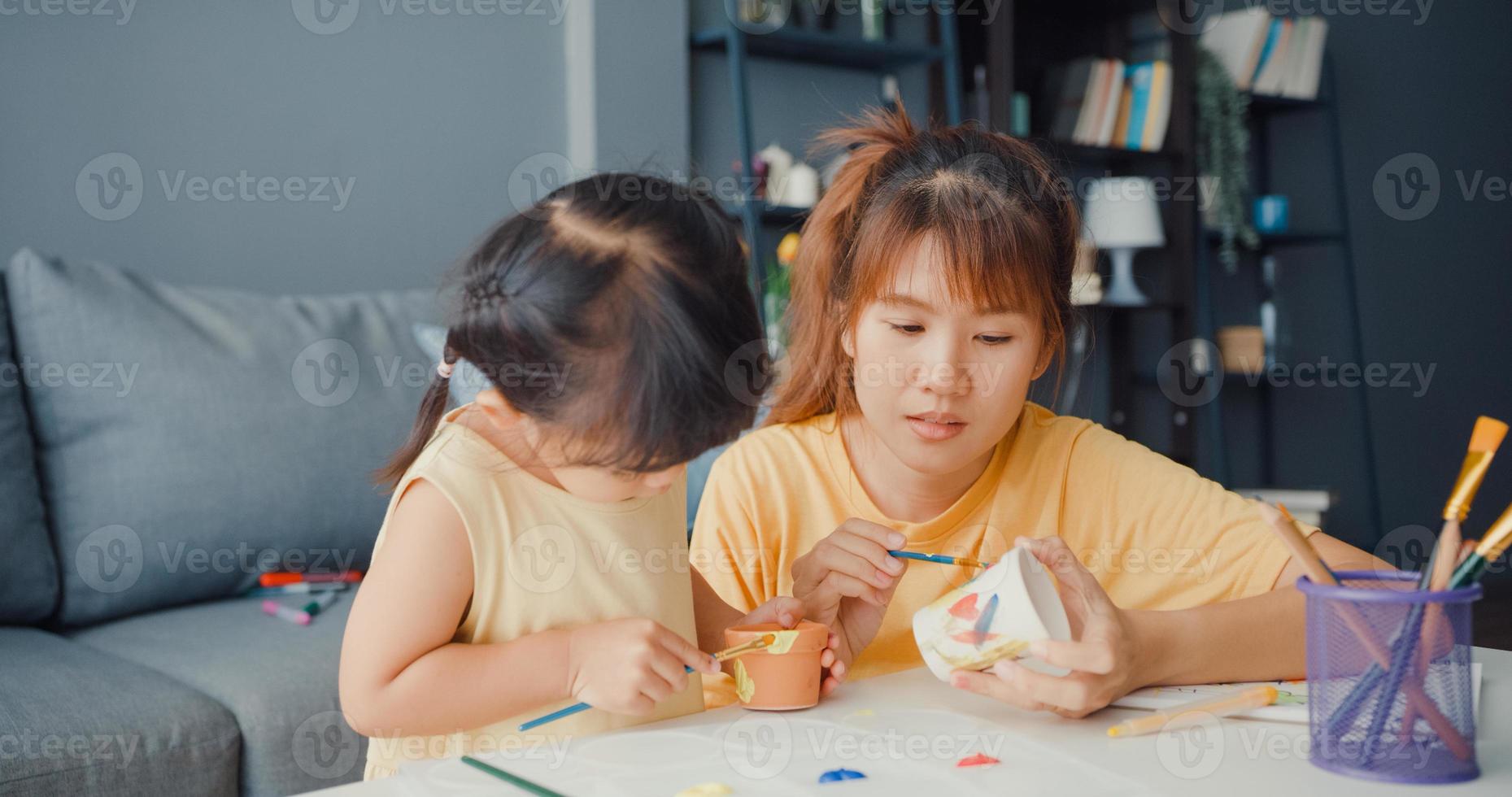 gelukkige vrolijke azië familie moeder leert peuter meisje verf keramische pot plezier ontspannen op tafel in de woonkamer thuis. tijd samen doorbrengen, sociale afstand, quarantaine voor coronaviruspreventie. foto