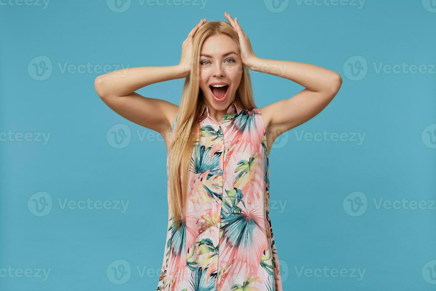 portret van versteld staan jong blond vrouw met gewoontjes kapsel poseren over- blauw achtergrond, houden palmen Aan haar hoofd en op zoek vrolijk Bij camera met breed mond geopend foto