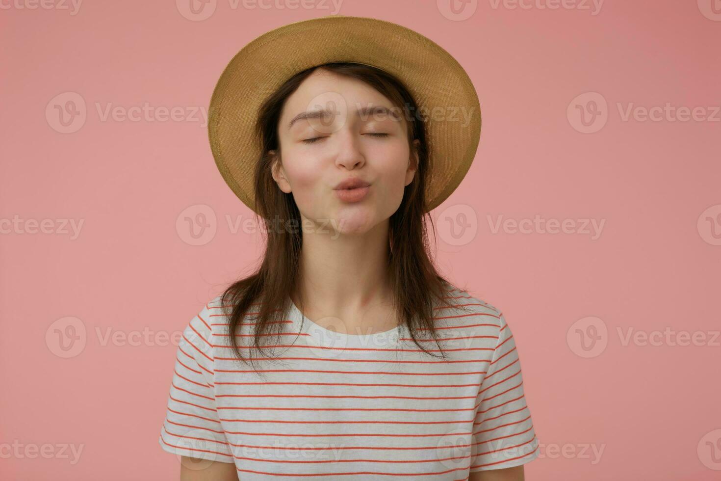 mooi hoor op zoek vrouw, mooi meisje met lang brunette haar. vervelend t-shirt met rood stroken en hoed. zoenen u met Gesloten ogen. emotioneel concept. staan geïsoleerd over- pastel roze achtergrond foto