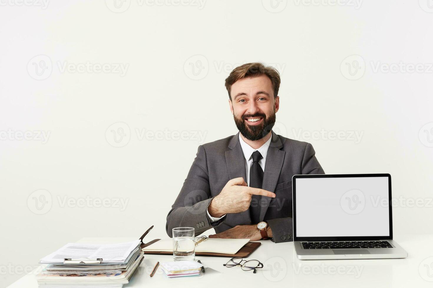 studio schot van brunette gebaard vent met kort kapsel poseren over- wit achtergrond in formeel kleren, tonen Aan zijn laptop met verheven hand- en op zoek vrolijk Bij camera foto