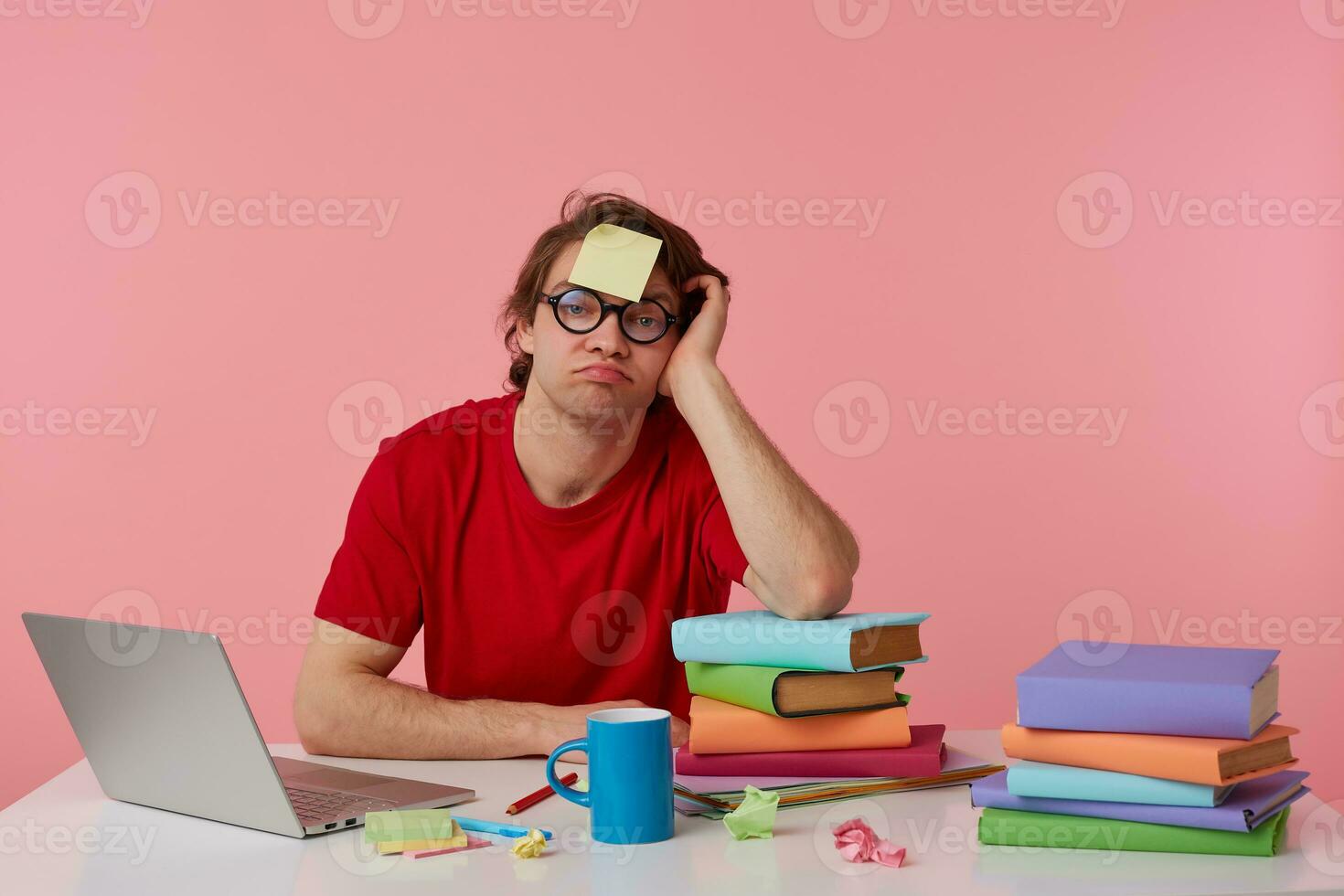 jong moe Mens in bril draagt in rood t-shirt, zit door de tafel en werken met notitieboekje en boeken, met een sticker Aan zijn voorhoofd, helaas looks Bij de camera, geïsoleerd over- roze achtergrond. foto