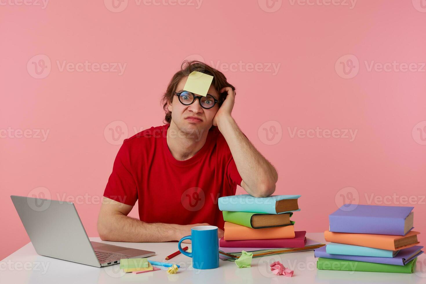 jong Mens in bril draagt in rood t-shirt, met een sticker Aan zijn voorhoofd, Mens zit door de tafel en werken met laptop en boeken, geïsoleerd over- roze achtergrond. looks niet tevreden en ongelukkig. foto