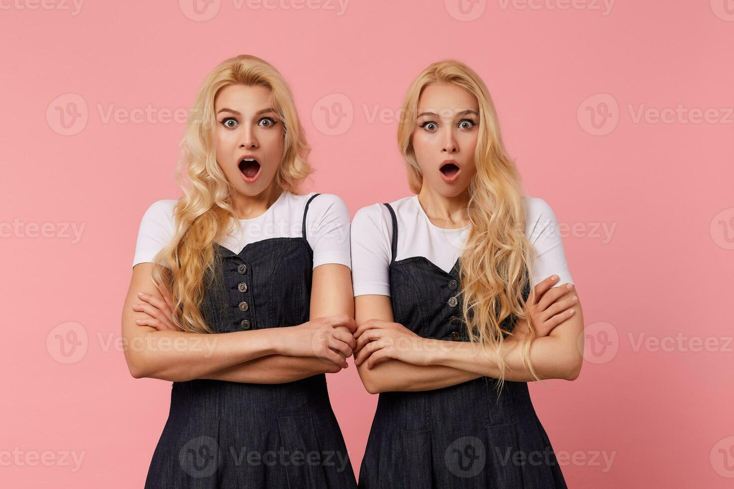 verbijsterd jong mooi lang haren blond Dames gekleed in elegant kleren houden handen gevouwen terwijl op zoek verbaasd Bij camera met breed ogen geopend, poseren over- roze achtergrond foto