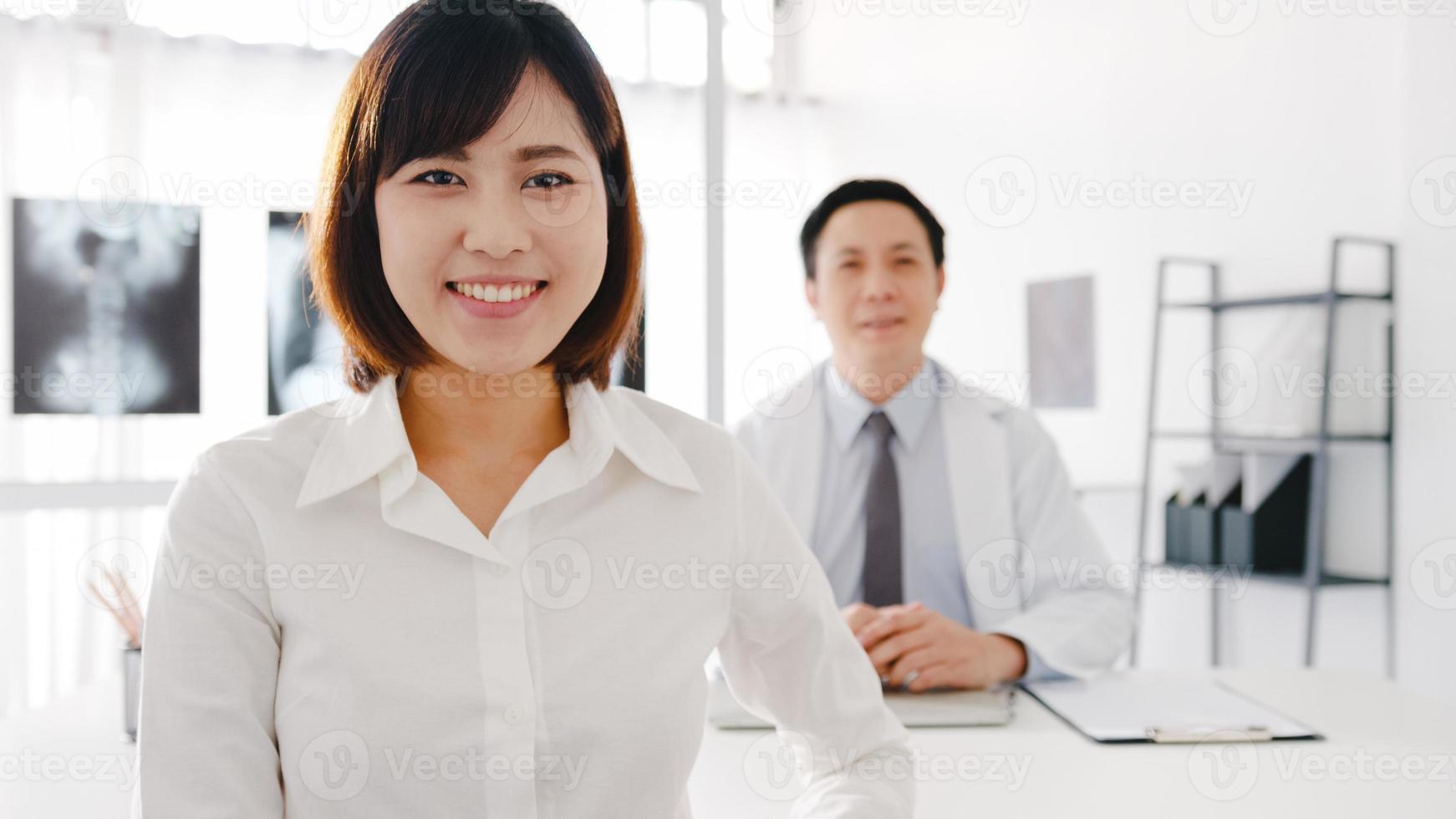 zelfverzekerde Aziatische mannelijke arts in wit medisch uniform en jong geduldig meisje dat naar de camera kijkt en glimlacht tijdens medisch consult aan de balie in de gezondheidskliniek of het ziekenhuis. advies- en therapieconcept foto