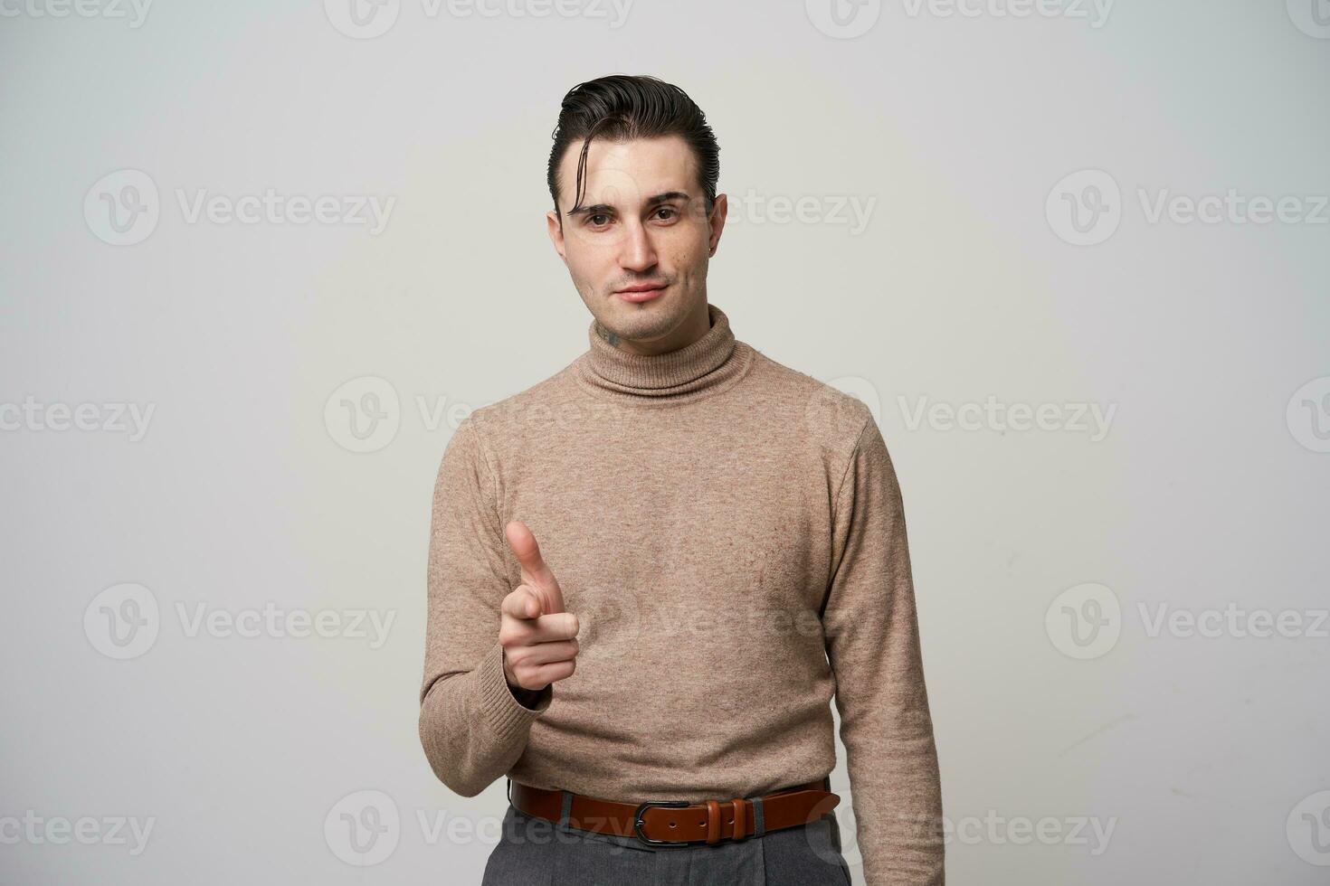 knap jong brunette mannetje hipster met elegant kapsel verhogen hand- en tonen Aan camera met wijsvinger, houden lippen gevouwen terwijl poseren over- wit achtergrond foto