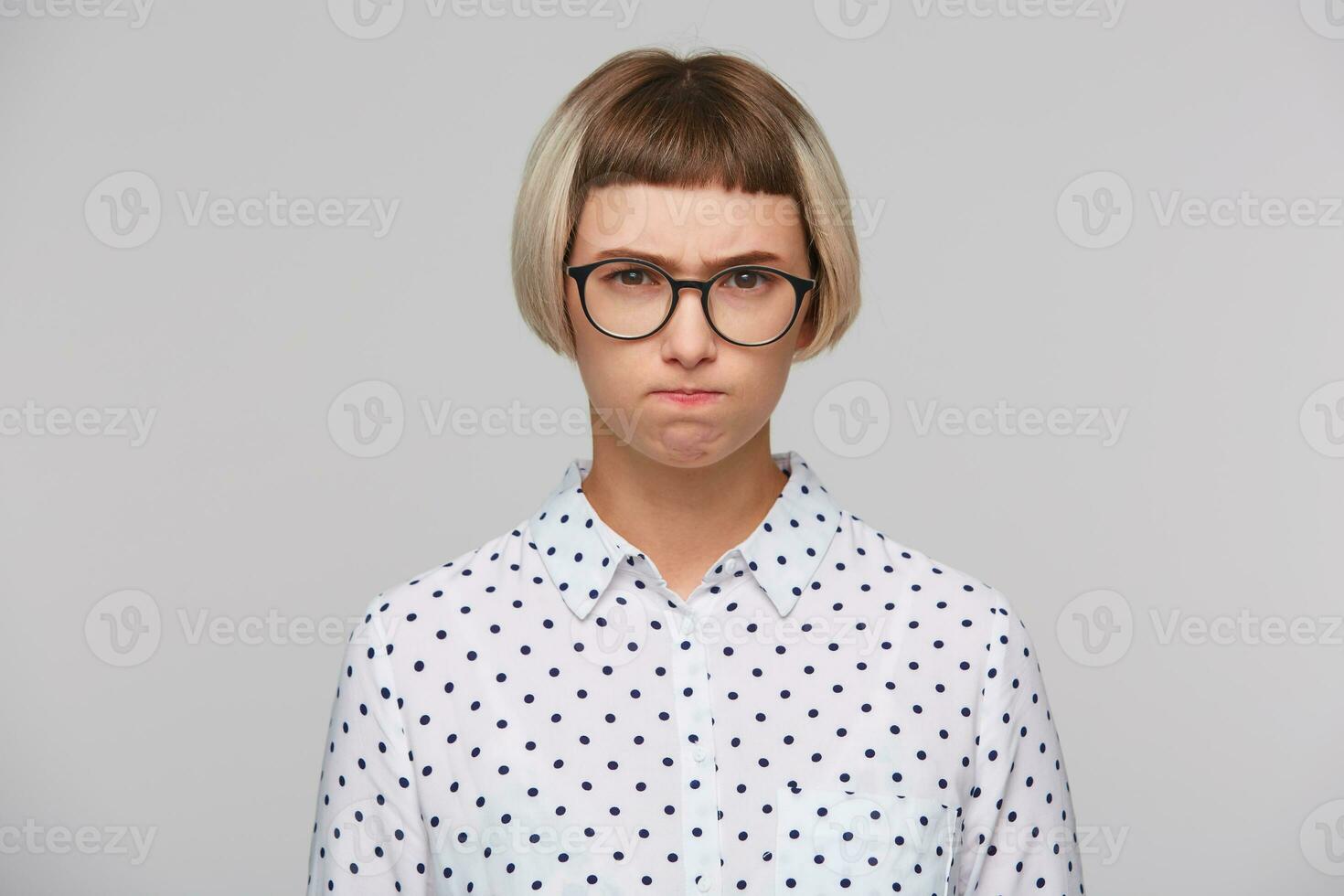 portret van echt geërgerd blond jong vrouw draagt polka punt overhemd en bril looks van streek en drukken lippen geïsoleerd over- wit achtergrond foto
