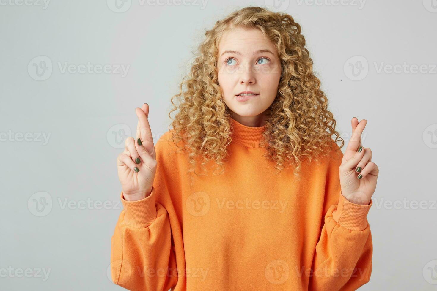 krullend haar blond in een helder oranje trui looks in de bovenste links hoek houdt vingers gekruiste bijten haar lip maken een wens, vraagt god voor helpen, hoopt voor haar geluk foto