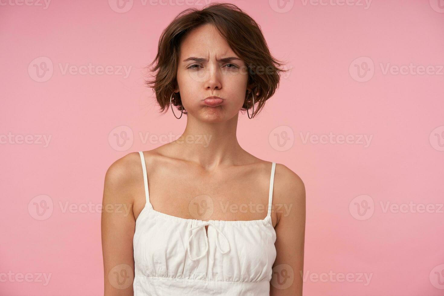 beledigd brunette vrouw met gewoontjes kapsel achtervolgen lippen en op zoek Bij camera met scheel, vervelend wit top terwijl staand over- roze achtergrond met handen naar beneden foto