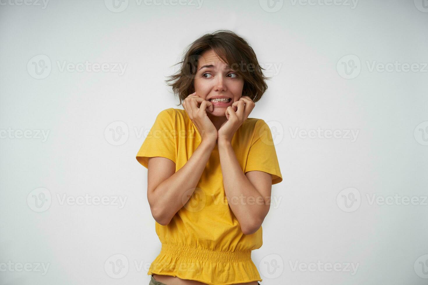 portret van bang jong vrouw met bruin haar- vervelend geel t-shirt, op zoek terzijde bang en Holding haar gezicht met verheven handen, geïsoleerd over- wit achtergrond foto