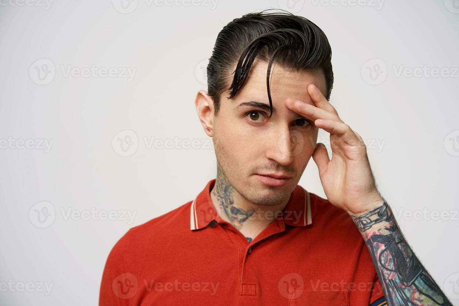 portret van een kalm, zelfverzekerd jongen, loensen Bij de camera, bruin ogen, intrigerend Look, zwart haar, styling, een arm verheven naar de gezicht, in een rood polo t-shirt, heeft tatoeages, Aan een wit achtergrond foto