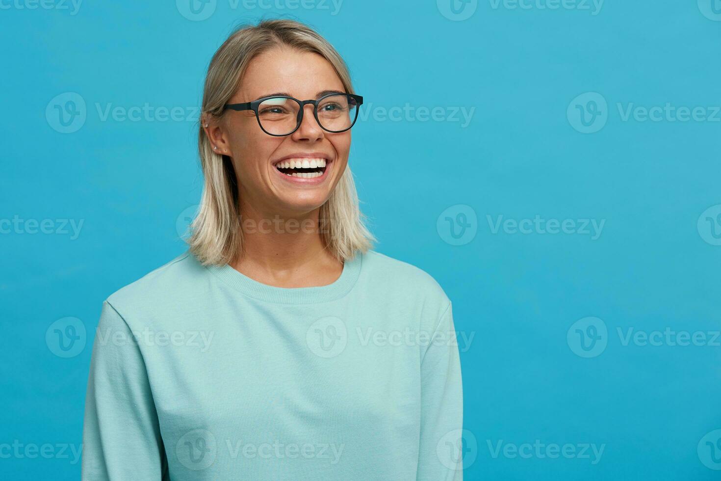 dichtbij omhoog van grappig vrolijk blij blond jong meisje in bril, toothy glimlach, lachend, staand zijwaarts, merkbaar gelukkig en optimistisch, gekleed in licht blauw lang mouw T-shirt, aan een blauw achtergrond foto