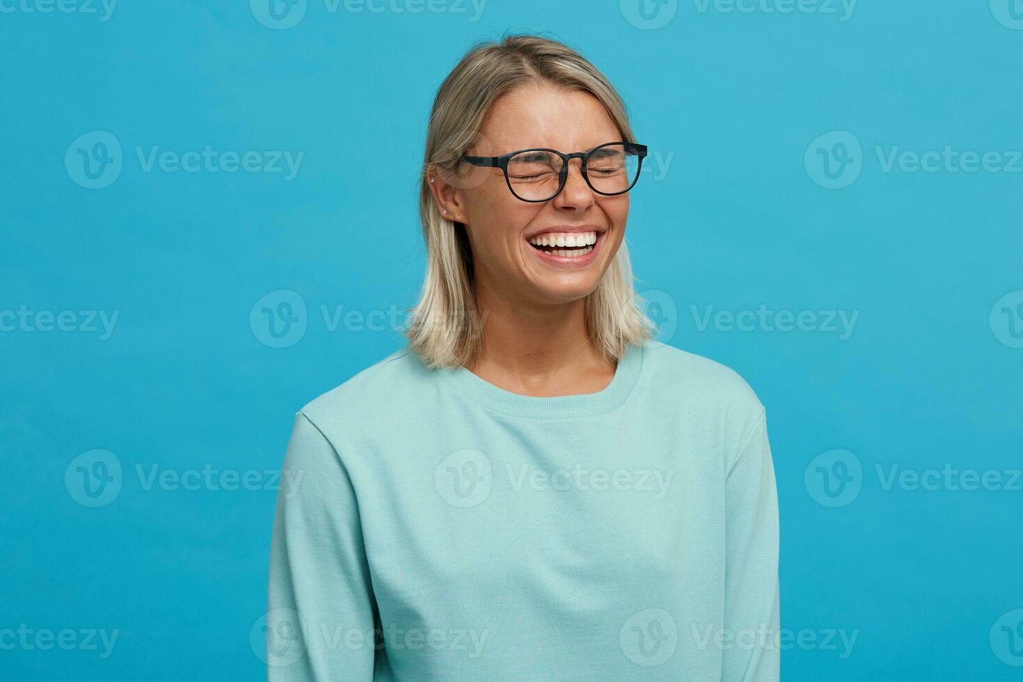 binnen- schieten van grappig vrolijk blij blond jong meisje in bril, breed glimlacht, sluit ogen terwijl lachend, staand zijwaarts, gekleed in licht blauw lang mouw T-shirt, aan een blauw achtergrond foto