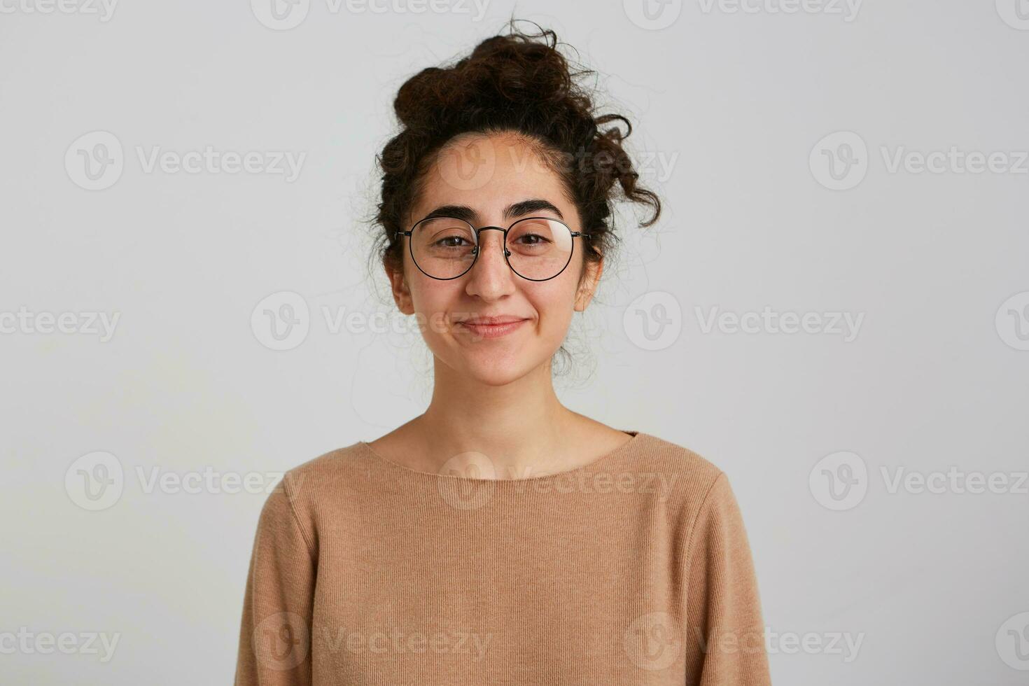 detailopname van glimlachen lief jong vrouw met bun van donker gekruld haar- draagt beige sweater en bril voelt gelukkig en tevreden geïsoleerd over- wit achtergrond looks direct in camera foto