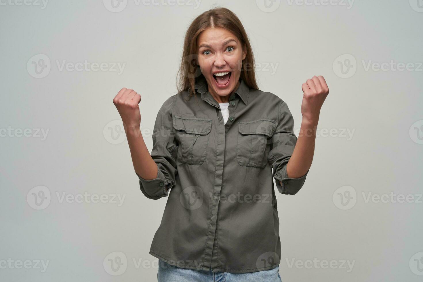 jong dame, mooi vrouw met bruin lang haar. vervelend grijs shirt, jeans en hijs- haar vuisten, gelukkig met succes, opgewonden. aan het kijken Bij de camera geïsoleerd over- grijs achtergrond foto