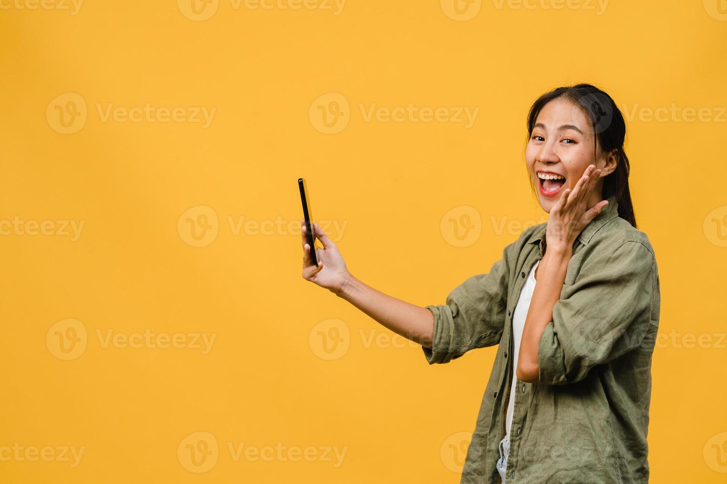 verraste jonge aziatische dame met behulp van mobiele telefoon met positieve uitdrukking, glimlacht breed, gekleed in casual kleding en staat geïsoleerd op gele achtergrond. gelukkige schattige blije vrouw verheugt zich over succes. foto