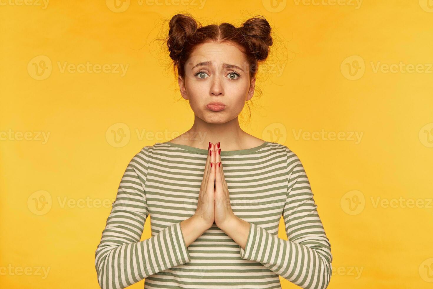 portret van aantrekkelijk, rood haar- meisje met twee broodjes. vervelend gestreept trui en aan het kijken verdrietig Bij de camera. vragen voor helpen, smeekt voor vergevingsgezind. staan geïsoleerd over- geel achtergrond foto