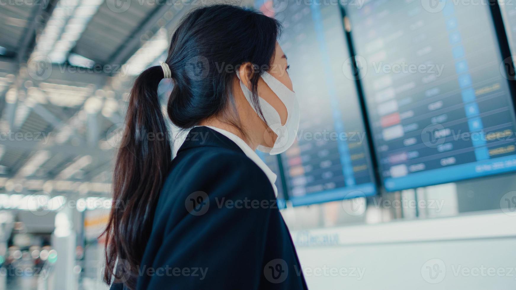 aziatisch zakenmeisje draagt een gezichtsmasker met een kofferstandaard voor het bord, kijk naar informatie die haar vlucht controleert op de internationale luchthaven. zakelijke forens covid pandemie, zakelijk reisconcept. foto