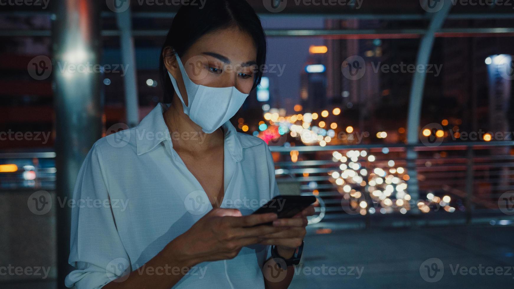 jonge azië zakenvrouw in mode kleding met gezichtsmasker met behulp van smartphone sms-bericht te typen terwijl je 's nachts buiten in de stedelijke stad staat. sociale afstand om verspreiding van het covid-19-concept te voorkomen. foto