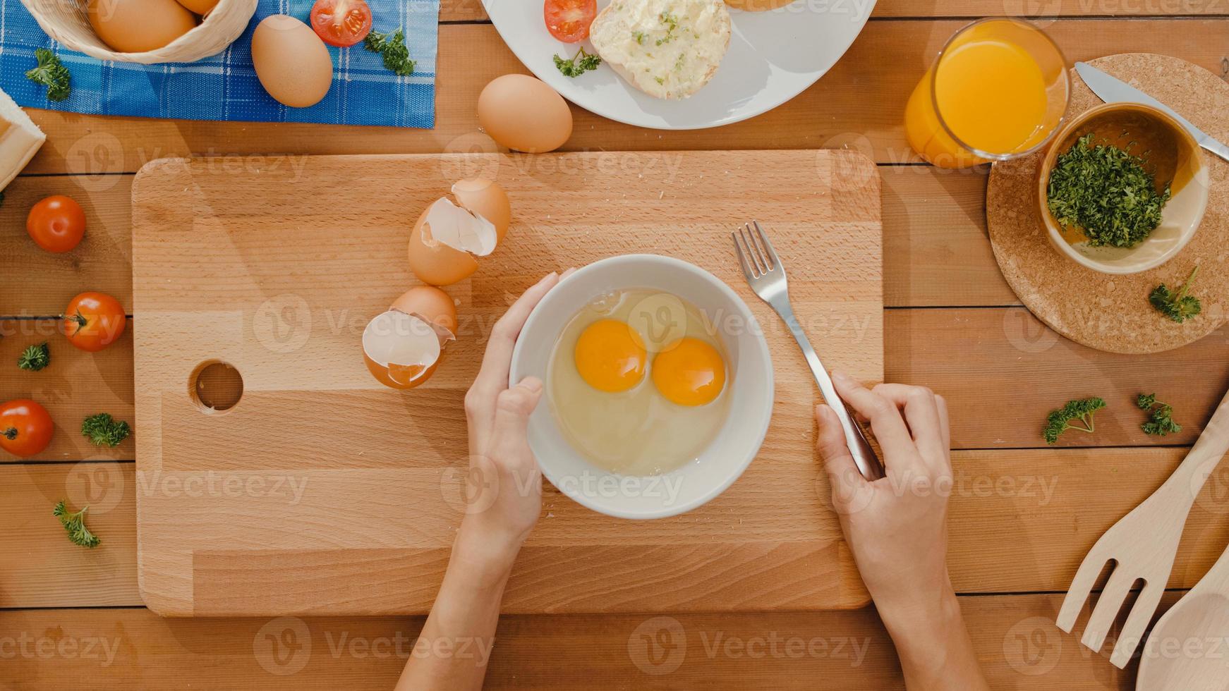 handen van jonge aziatische vrouw chef-kok die ei in keramische kom zwaait, kook omelet met groenten op een houten bord op de keukentafel in huis. levensstijl gezond eten en traditionele bakkerij. bovenaanzicht schot. foto