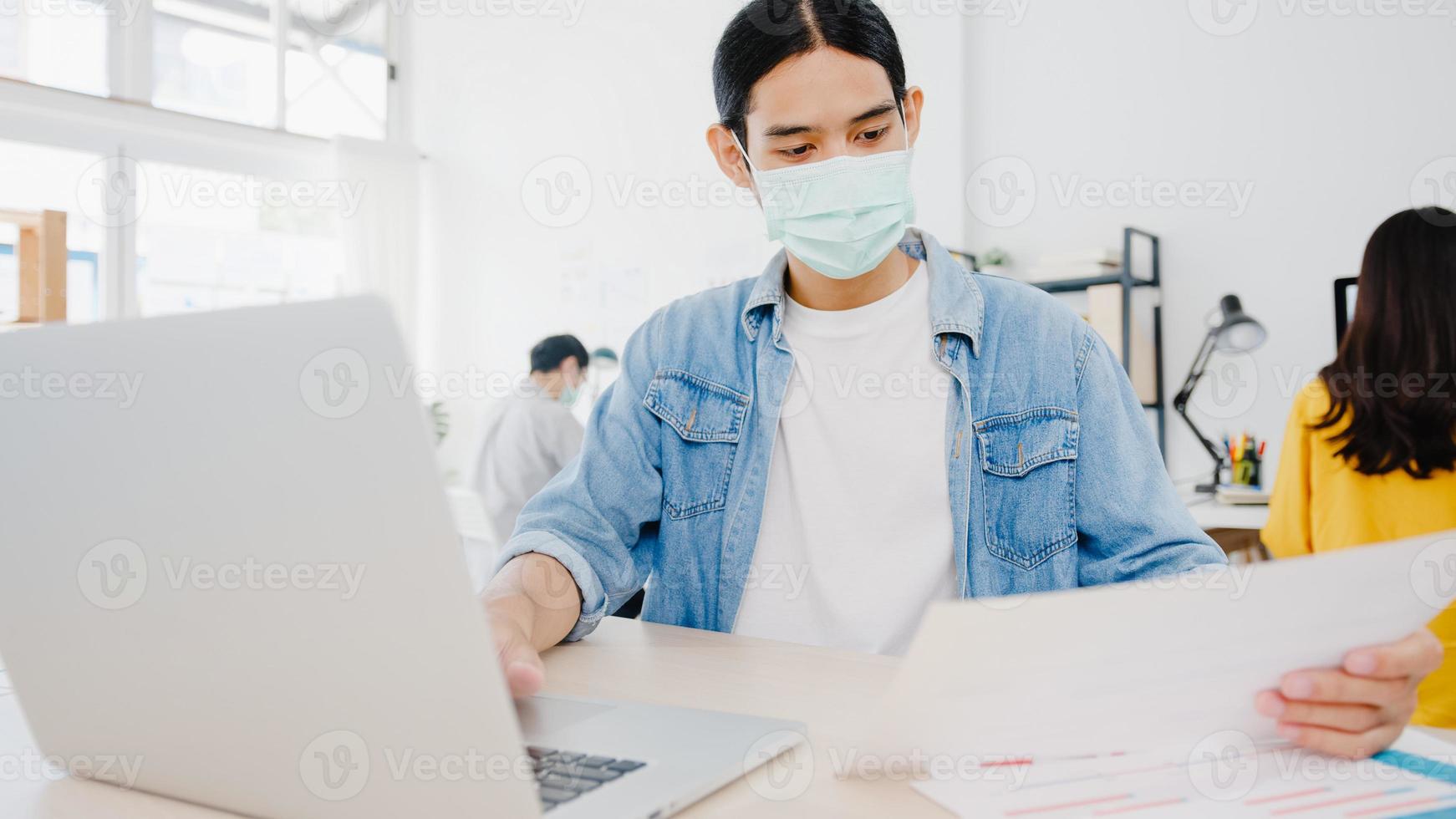 azische zakenman-ondernemer die een medisch gezichtsmasker draagt voor sociale afstand in een nieuwe normale situatie voor viruspreventie tijdens het gebruik van een laptop op het werk op kantoor. levensstijl na het coronavirus. foto