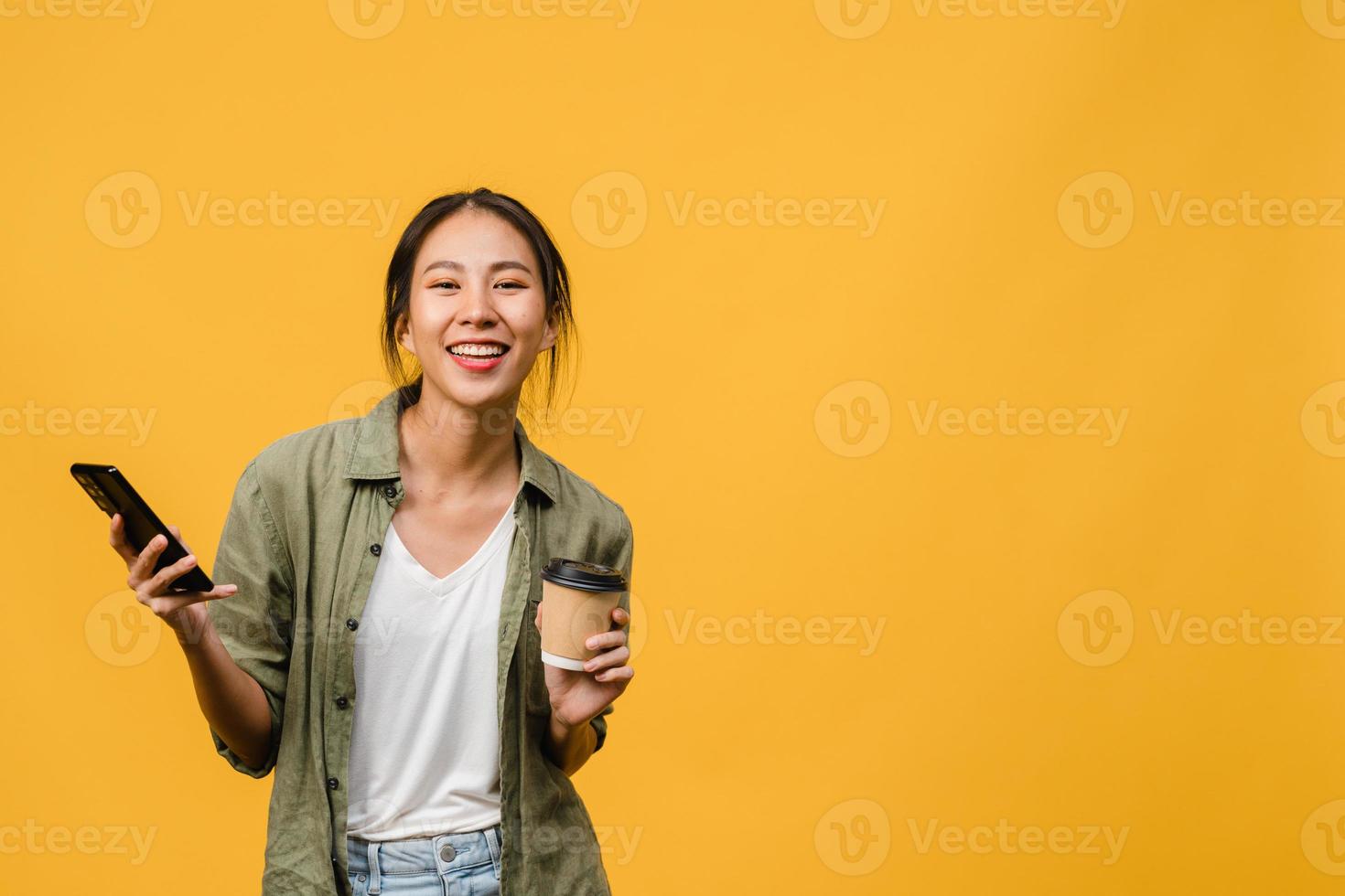verraste jonge aziatische dame die telefoon gebruikt en koffiekopje vasthoudt met positieve uitdrukking, breed glimlacht, gekleed in casual kleding en kijkt naar camera op gele achtergrond. gezichtsuitdrukking concept. foto