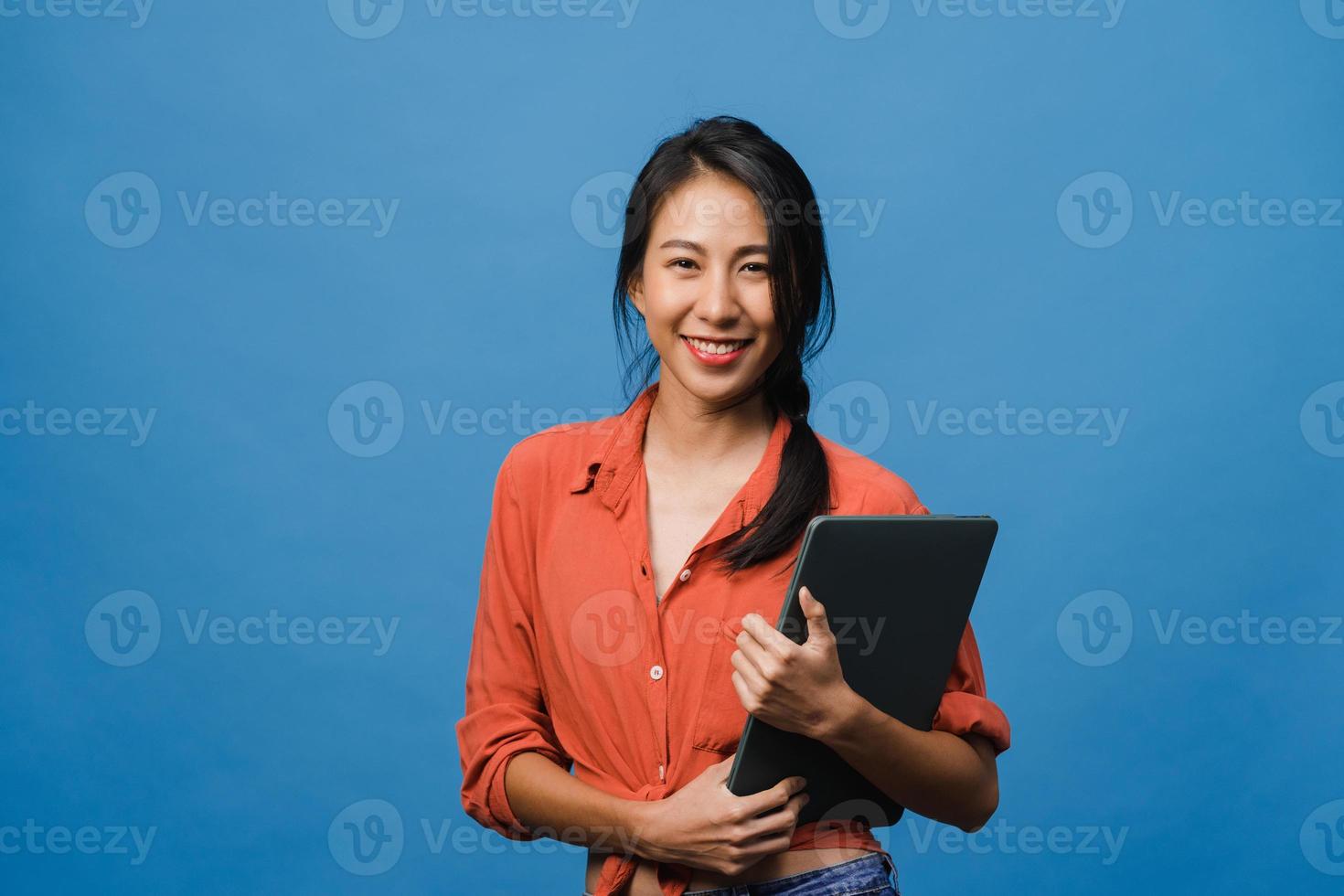 verraste jonge aziatische dame houdt laptop vast met positieve uitdrukking, glimlach breed, gekleed in casual kleding en kijkt naar camera op blauwe achtergrond. gelukkige schattige blije vrouw verheugt zich over succes. foto