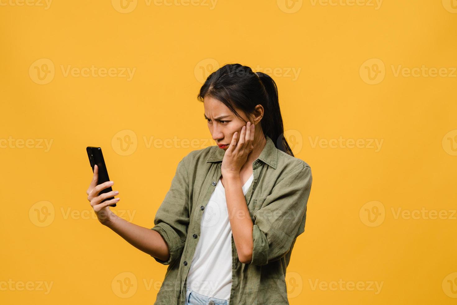 denken dromende jonge azië dame met behulp van telefoon met positieve uitdrukking, gekleed in casual doek gevoel van geluk en staan geïsoleerd op gele achtergrond. gelukkige schattige blije vrouw verheugt zich over succes. foto