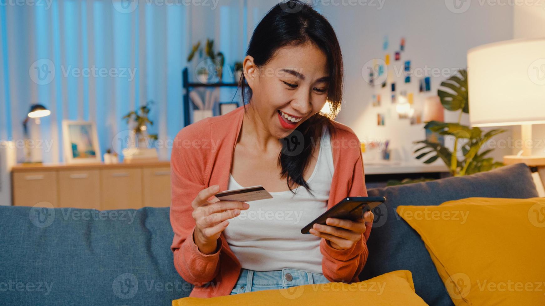 's nachts mooie lachende aziatische dame gebruik mobiel om online winkelproduct met creditcard op de bank in de woonkamer te bestellen. blijf thuis, zelf quarantaine activiteit, leuke activiteit voor covid quarantaine. foto