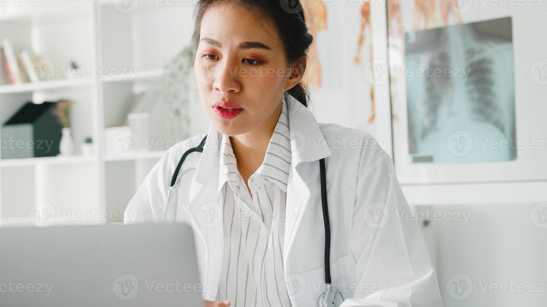 jonge Aziatische dame arts in wit medisch uniform met stethoscoop met behulp van computerlaptop praten videoconferentiegesprek met patiënt aan balie in gezondheidskliniek of ziekenhuis. advies- en therapieconcept. foto