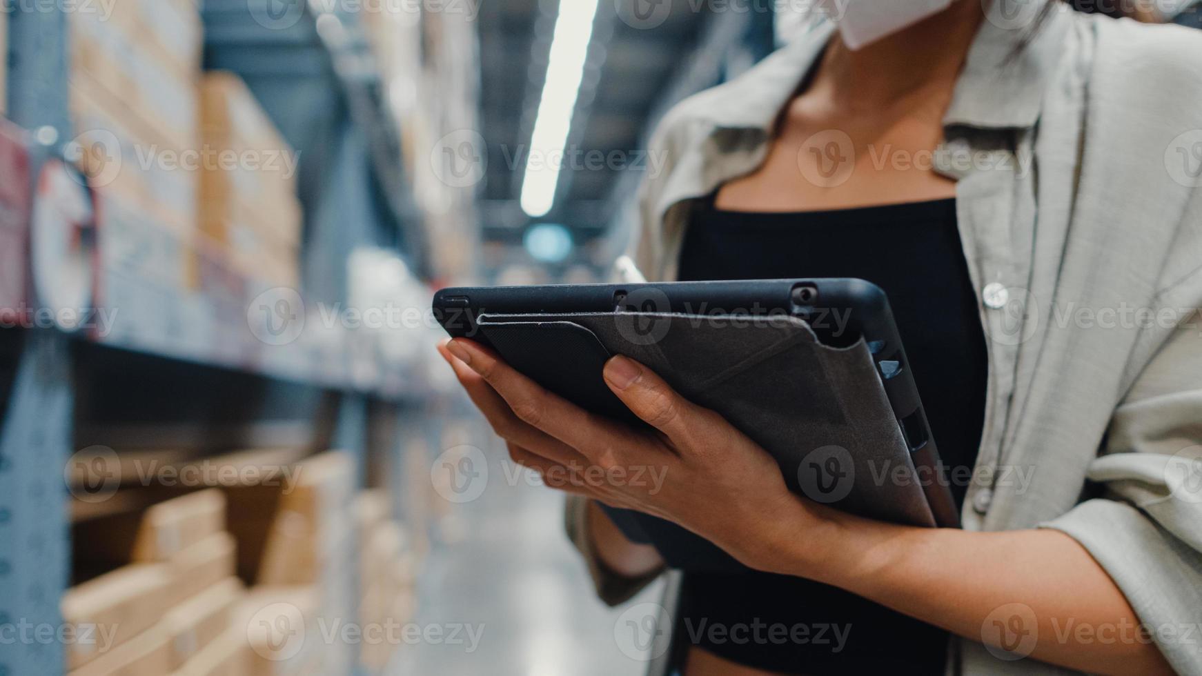 jonge azië zakenvrouw manager draagt gezichtsmasker op zoek naar goederen met behulp van digitale tablet controleren voorraadniveaus staan in retail winkelcentrum. distributie, logistiek, pakketten klaar voor verzending. foto