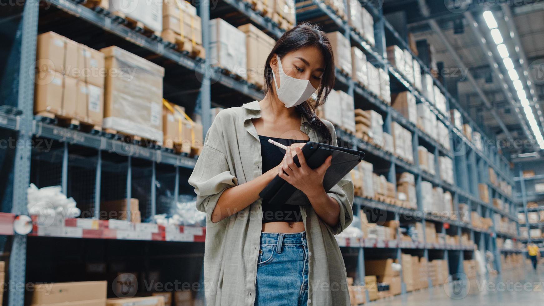 jonge azië zakenvrouw manager draagt gezichtsmasker op zoek naar goederen met behulp van digitale tablet controleren voorraadniveaus staan in retail winkelcentrum. distributie, logistiek, pakketten klaar voor verzending. foto