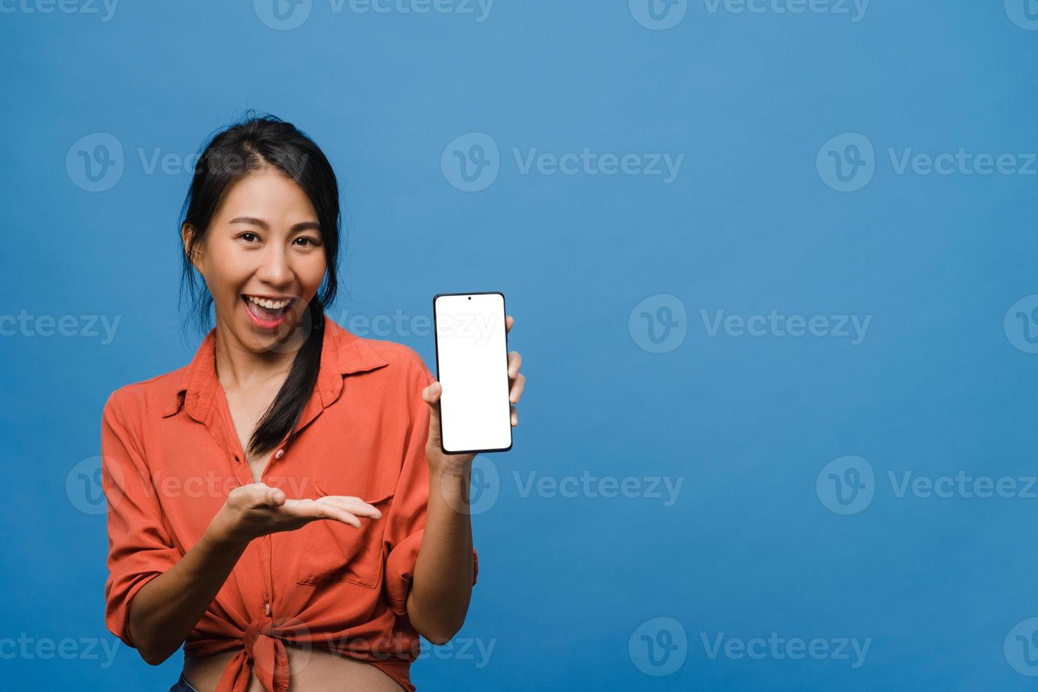 jonge aziatische dame toont een leeg smartphonescherm met positieve uitdrukking, glimlacht breed, gekleed in casual kleding en voelt zich gelukkig op een blauwe achtergrond. mobiele telefoon met wit scherm in vrouwelijke hand. foto