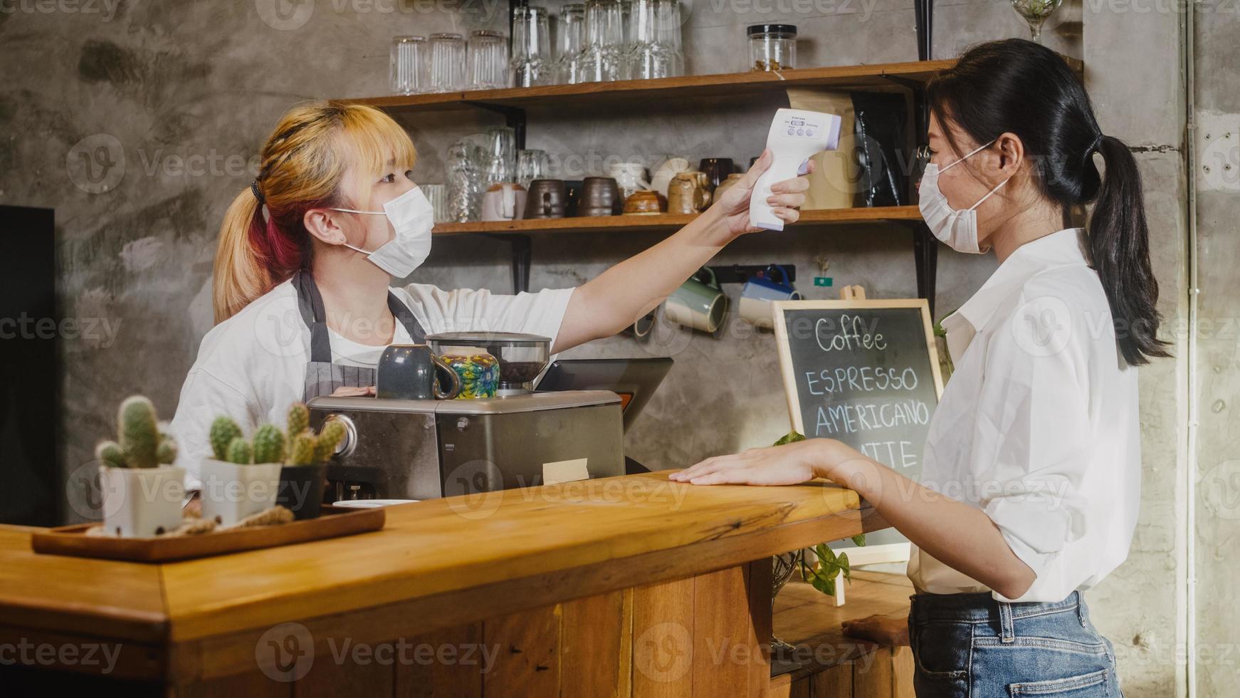 jonge Aziatische vrouwelijke restaurantmedewerkers die een beschermend gezichtsmasker dragen met behulp van een infraroodthermometercontrole of een temperatuurpistool op het voorhoofd van de klant voordat ze naar binnen gaan. levensstijl nieuw normaal na corona virus. foto