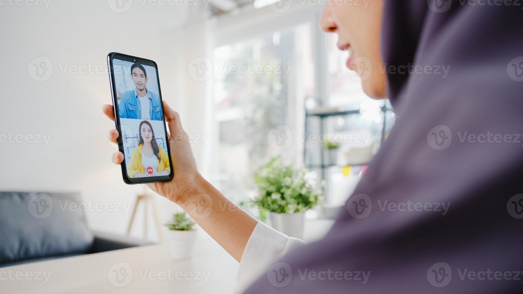 jonge azië moslim zakenvrouw met behulp van slimme telefoon praten met collega door videochat brainstorm online vergadering terwijl op afstand werken vanuit huis in de woonkamer. sociale afstand, quarantaine voor het coronavirus. foto