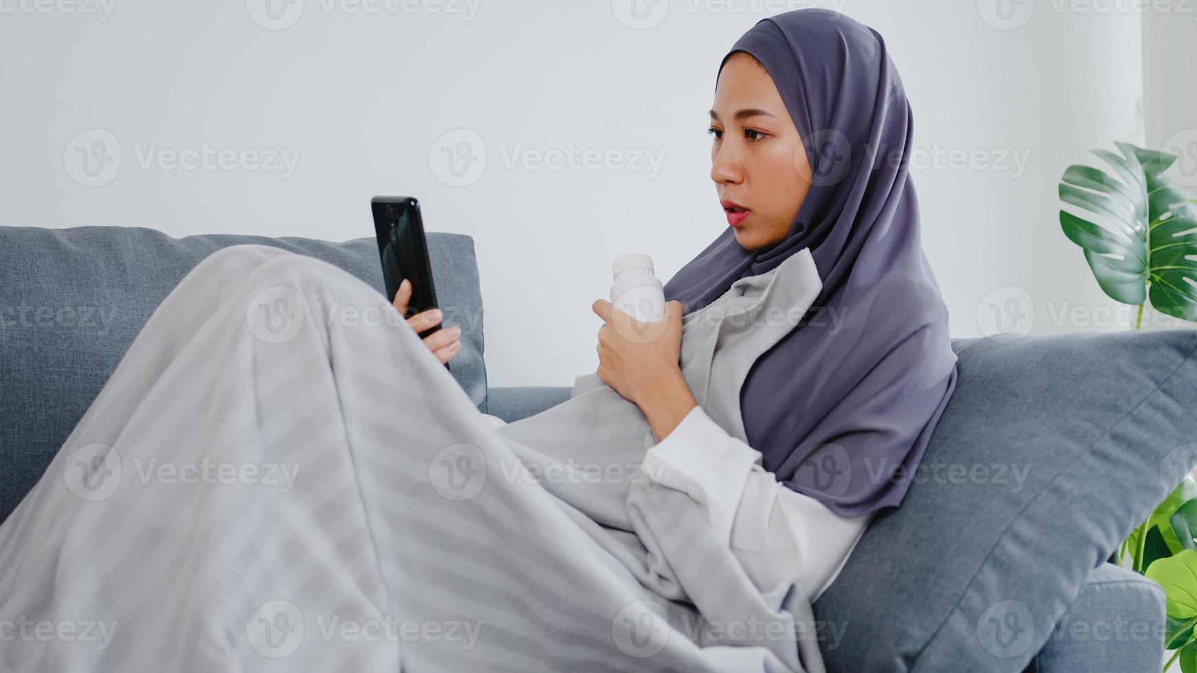 jonge azische moslimdame draagt hijab met behulp van een telefoonvideogesprek met een doktersconsultatie of online consultatie op de bank in de woonkamer thuis. sociale afstand, quarantaine voor coronavirusconcept. foto