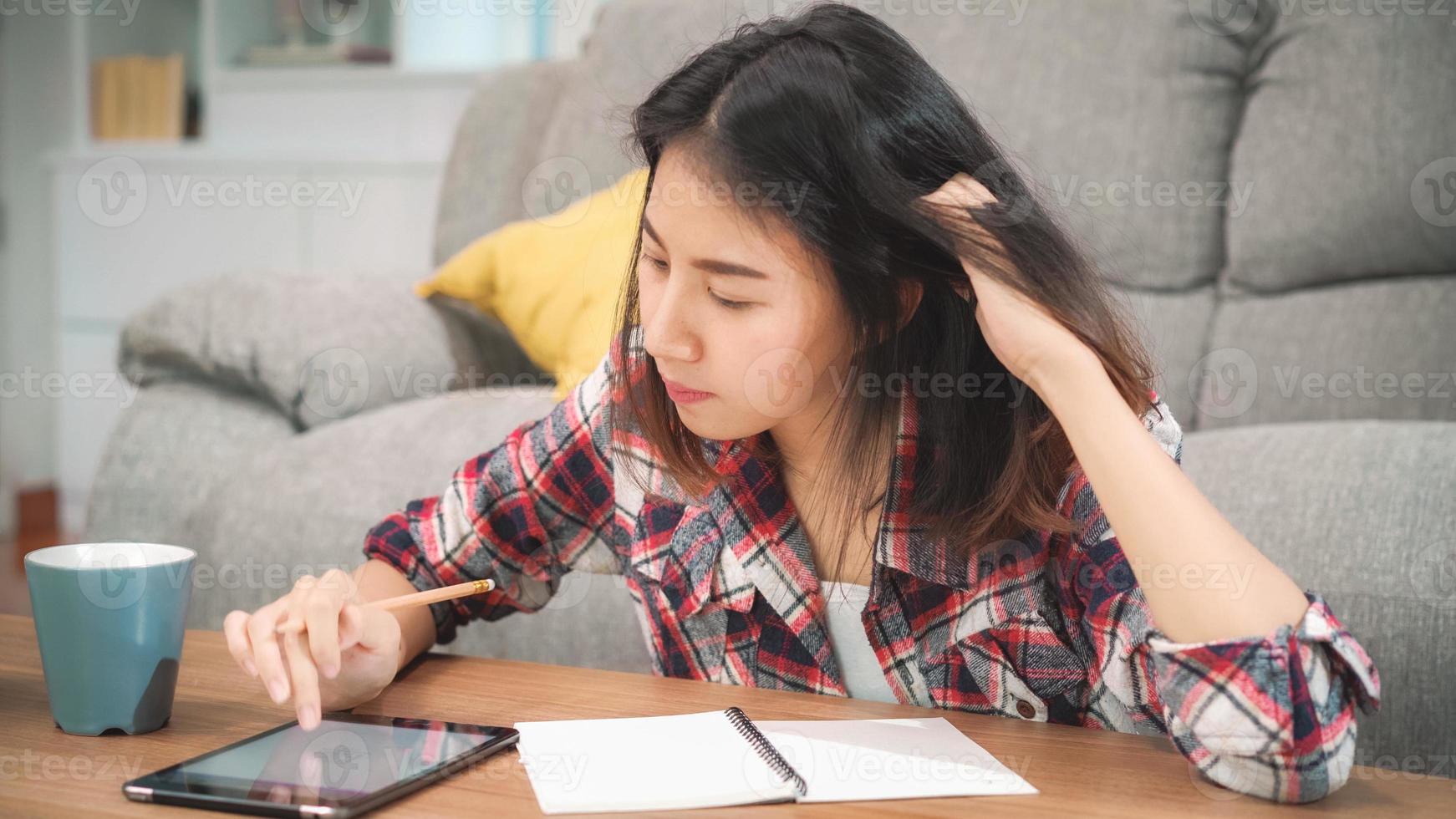 Aziatische studentenvrouw doet thuis huiswerk, vrouw drinkt koffie en gebruikt een tablet om thuis op de bank in de woonkamer te zoeken. levensstijl vrouwen ontspannen thuis concept. foto
