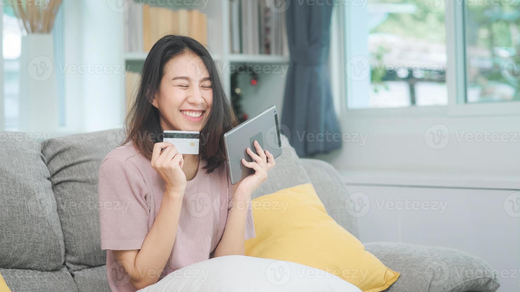 jonge glimlachende aziatische vrouw die tablet gebruikt om online winkelen met een creditcard te kopen terwijl ze op de bank ligt wanneer ze thuis in de woonkamer ontspant. levensstijl Latijnse en Spaanse etniciteit vrouwen bij huis concept. foto