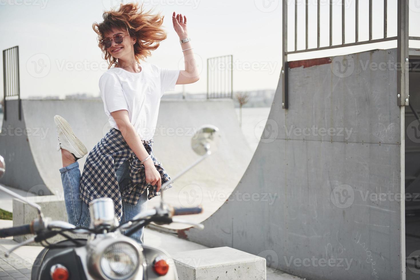 portret van een mooi meisje hipster zittend op een zwarte retro scooter, glimlachend poseren en genieten van de warme lentezon. foto