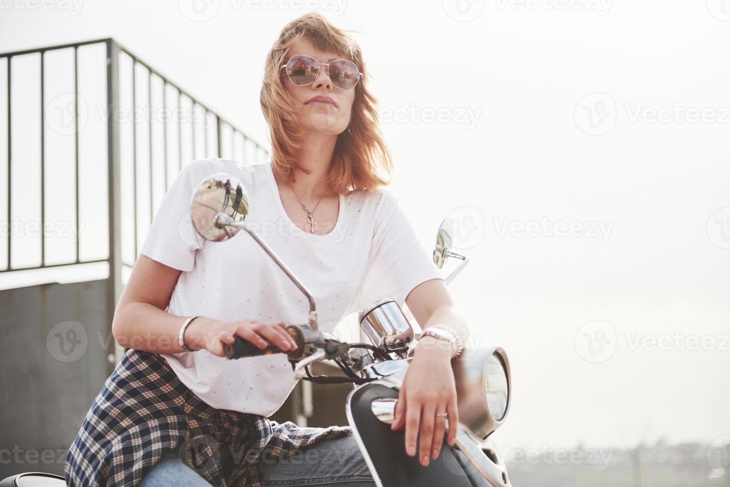 portret van een mooi meisje hipster zittend op een zwarte retro scooter, glimlachend poseren en genieten van de warme lentezon. foto