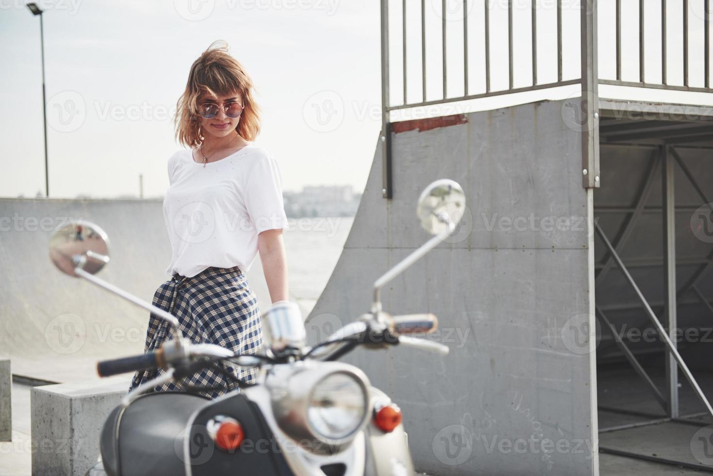 portret van een mooi meisje hipster zittend op een zwarte retro scooter, glimlachend poseren en genieten van de warme lentezon. foto