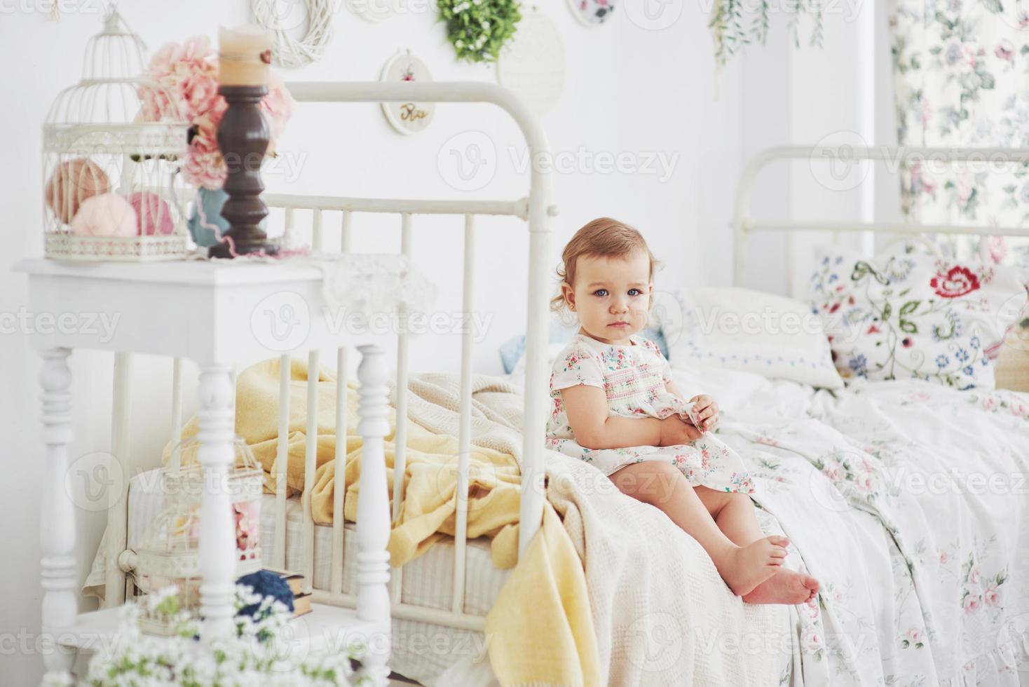 kindertijd concept. meisje van de baby in schattige jurk aanbrengen op bed spelen met speelgoed door het huis. witte vintage kinderkamer foto