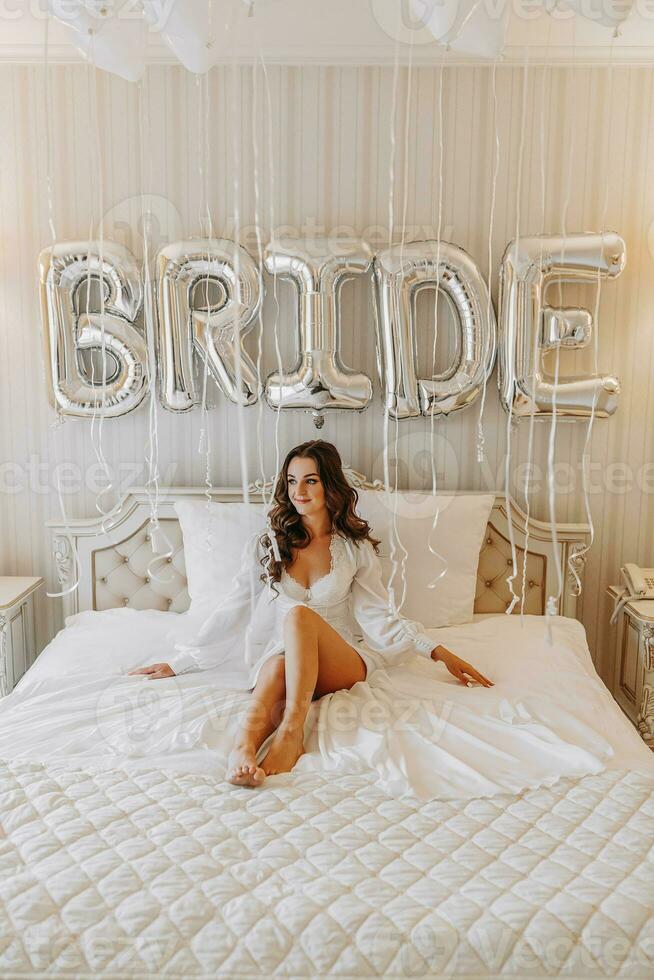 jong gekruld meisje met lang haar- in luxueus kamer zittend Aan bed in wit Open jurk en voorbereidingen treffen voor bruiloft ceremonie foto