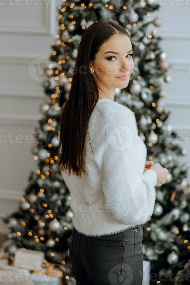 een mooi en elegant jong meisje in een wit trui poses in de Kerstmis lichten Aan de achtergrond van de Kerstmis boom foto