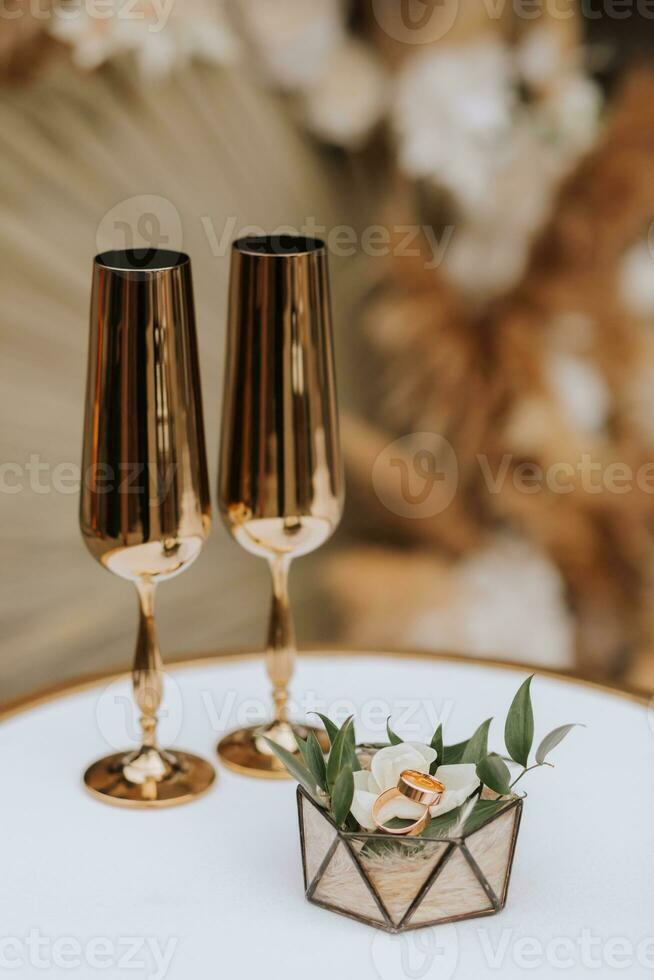 goud bruiloft ringen Aan een glas doos versierd met roos bloemen, De volgende naar het zijn bruiloft bril in goud kleur foto