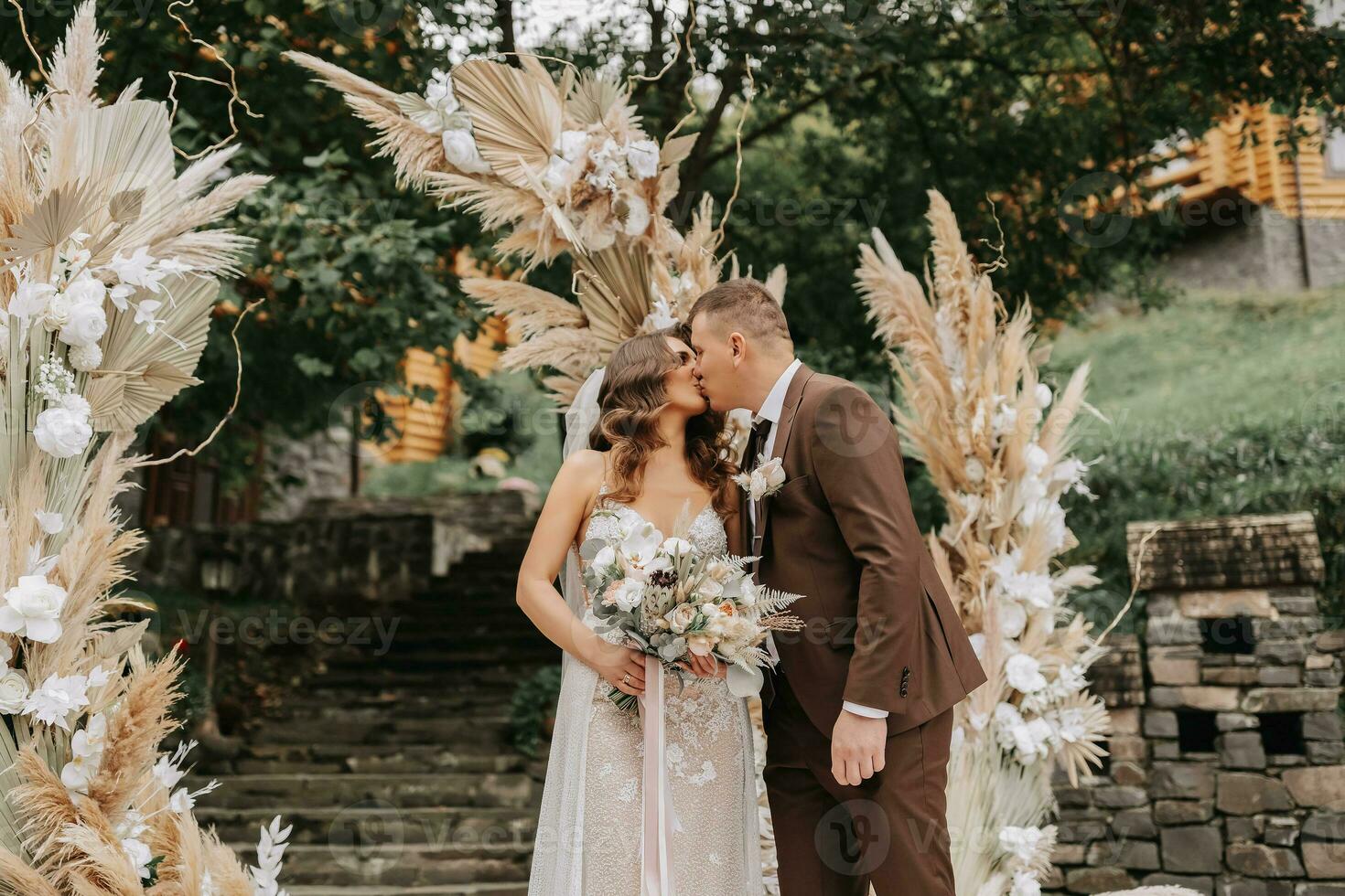 bruidegom en bruid kus. pasgetrouwden met een bruiloft boeket staand Bij een bruiloft ceremonie onder een boog versierd met bloemen en droog bloemen buitenshuis. foto