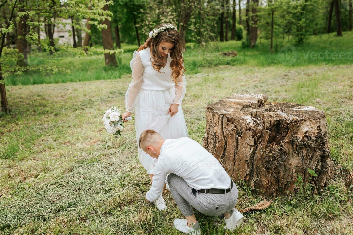 bruids wandelen in de Woud. de bruidegom is gekleed in een wit overhemd en grijs broek, helpen de bruid naar stropdas haar schoenveters foto