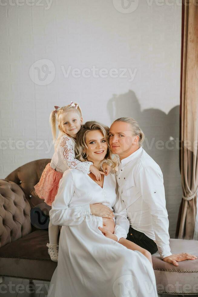 studio portret van een mooi zwanger jong vrouw in een wit jurk met een trein zittend Aan een sofa met haar weinig dochter en haar man foto